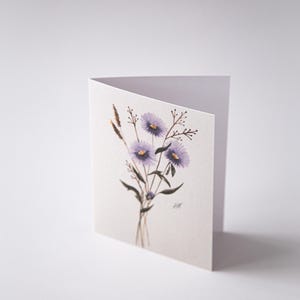 Può includere: Biglietto di auguri piegato con un'illustrazione ad acquerello di fiori viola e rametti in un vaso trasparente. Il biglietto è bianco con il disegno floreale sul davanti. Le iniziali dell'artista, "HM", sono in basso a destra.