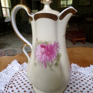May include: A white porcelain teapot with a gold trim and a floral design featuring pink chrysanthemums. The teapot has a lid with a gold knob and a handle.