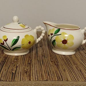 May include: A vintage cream and sugar set. The set includes a lidded sugar bowl and a creamer. Both pieces are white ceramic with yellow flower and green leaf designs. The handles are beaded. The sugar bowl has a round shape.