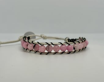 May include: A wrap bracelet featuring pink gemstone beads, black stitching, and a white leather band. The bracelet is secured with a white leather tie closure. The beads are approximately 0.3 inches in diameter.