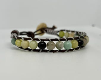 May include: A wrap bracelet featuring a series of colorful, round beads in shades of green, yellow, black, and cream. The beads are strung on a brown leather band, with a silver-colored thread woven between the beads. The bracelet is secured with a button closure.