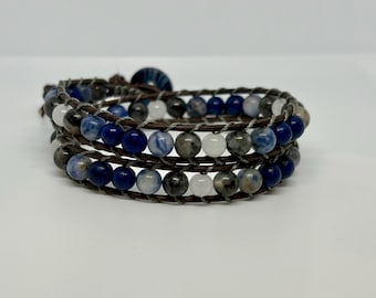 May include: Wrap bracelet featuring three strands of dark brown leather, adorned with blue, white, and gray gemstone beads. The bracelet is secured with a dark blue button clasp. A stylish accessory.