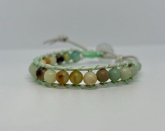 May include: Wrap bracelet featuring a row of colorful, round beads in shades of green, brown, and yellow. The beads are strung on a light gray leather band, accented with mint green thread. The bracelet is secured with a leather tie.