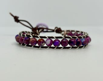 May include: Wrap bracelet featuring deep purple and maroon beads, strung on a brown leather band. The beads are interspersed with white thread, and the bracelet has a leather tie closure. The bracelet is a jewelry item.