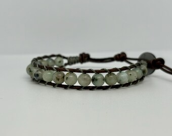 May include: A wrap bracelet featuring light green and speckled beads. The beads are strung on a dark brown leather cord, with a braided design between the beads. The bracelet has a button closure.