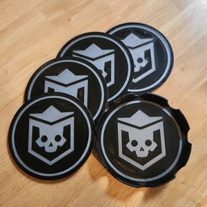 May include: Five black and silver round coasters with a skull and crown design. The coasters are made of a textured material and have a raised edge.