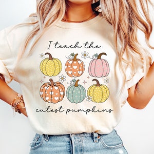 May include: Cream-colored t-shirt featuring a graphic of six pumpkins in various colors and patterns, with the text "I teach the cutest pumpkins." The pumpkins have heart, polka dot, and floral designs. A fall-themed shirt.