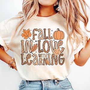 May include: A white t-shirt with the text "Fall in love with learning" in brown and orange lettering. The text is decorated with small hearts, leaves, and a pumpkin.