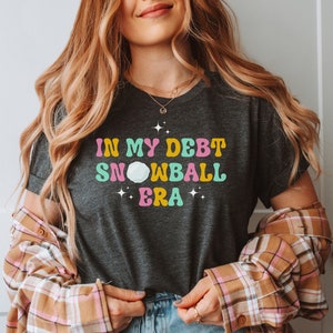 May include: A dark gray t-shirt with the text "In My Debt Snowball Era" in colorful retro lettering. The text is surrounded by small white stars.