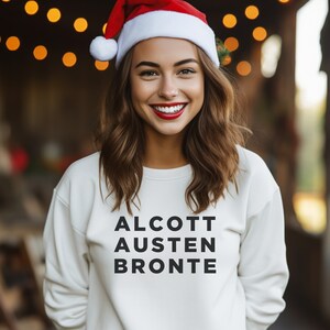 May include: A white sweatshirt with the words "ALCOTT AUSTEN BRONTE" printed in black on the front. The person wearing the sweatshirt is wearing a red Santa hat.