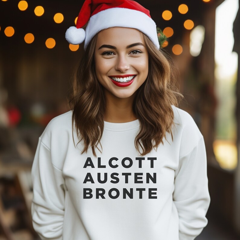 May include: A white sweatshirt with the words "ALCOTT AUSTEN BRONTE" printed in black on the front. The person wearing the sweatshirt is wearing a red Santa hat.