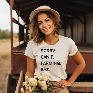 K&ouml;nnte beinhalten: Eine Frau tr&auml;gt ein wei&szlig;es T-Shirt mit dem Text "SORRY. CAN'T. FARMING. BYE." darauf. Sie h&auml;lt einen Strau&szlig; wei&szlig;er Blumen und steht vor einer Holzkonstruktion.