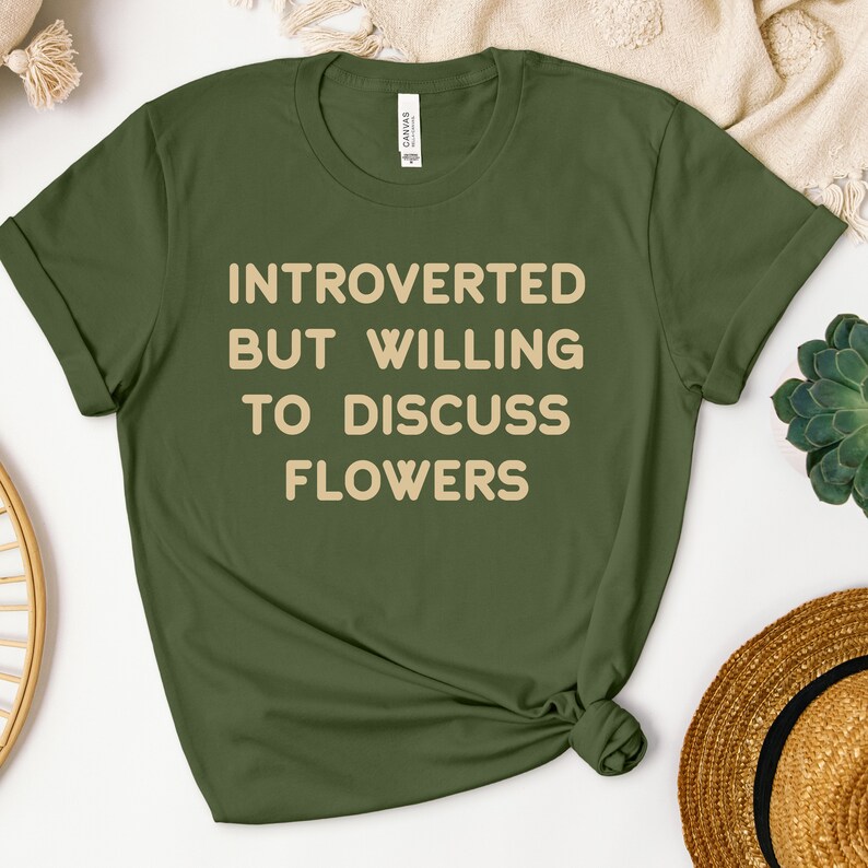 K&ouml;nnte beinhalten: Ein gr&uuml;nes T-Shirt mit dem Text "Introvertiert, aber bereit, &uuml;ber Blumen zu diskutieren" in hellbrauner Farbe.