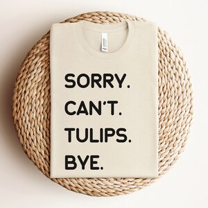 K&ouml;nnte beinhalten: Ein beiges T-Shirt mit schwarzem Text, der "SORRY. CAN'T. TULIPS. BYE." lautet.