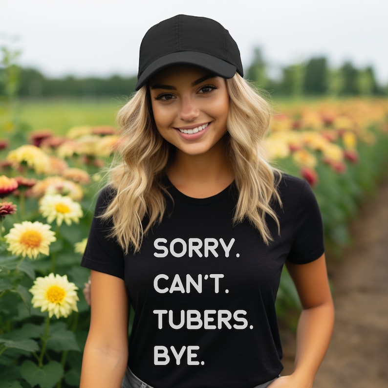K&ouml;nnte beinhalten: Eine Frau tr&auml;gt ein schwarzes T-Shirt mit dem Text "SORRY. CAN'T. TUBERS. BYE." in wei&szlig;en Buchstaben. Sie tr&auml;gt auch eine schwarze Baseballkappe und steht auf einem Feld mit Blumen.