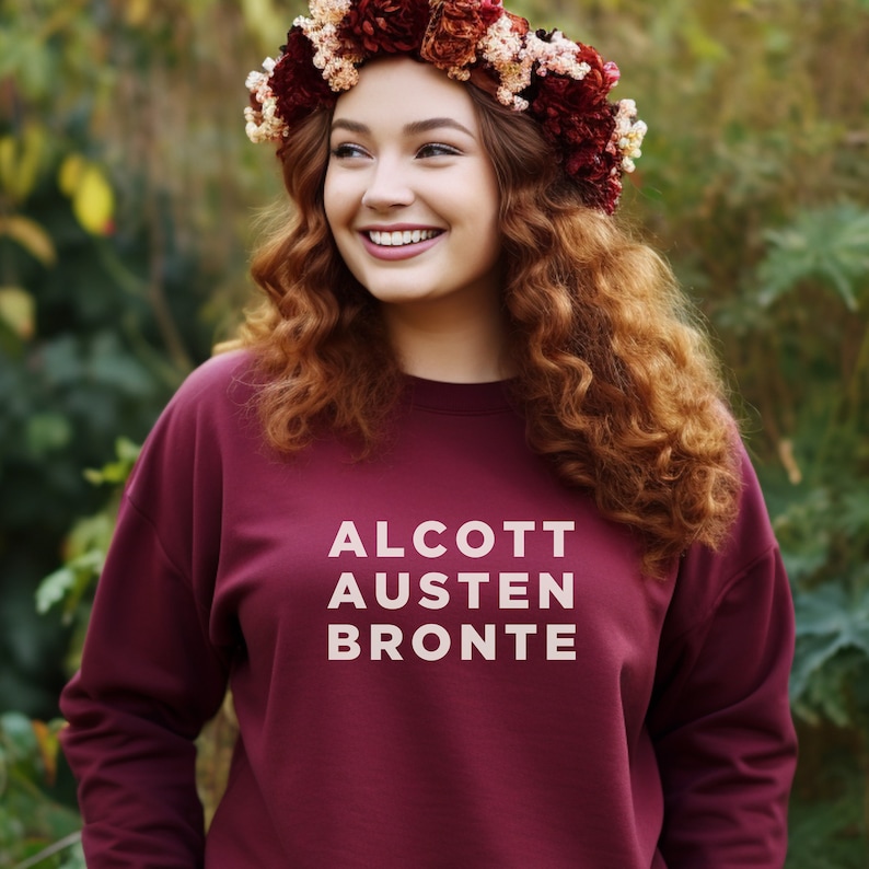 May include: A maroon sweatshirt with the words "ALCOTT AUSTEN BRONTE" printed in white on the front. The model is wearing a flower crown.