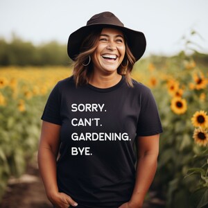 K&ouml;nnte beinhalten: Eine Frau tr&auml;gt ein schwarzes T-Shirt mit dem Text "SORRY. CAN'T. GARDENING. BYE." in wei&szlig;en Buchstaben. Sie l&auml;chelt und steht in einem Sonnenblumenfeld.