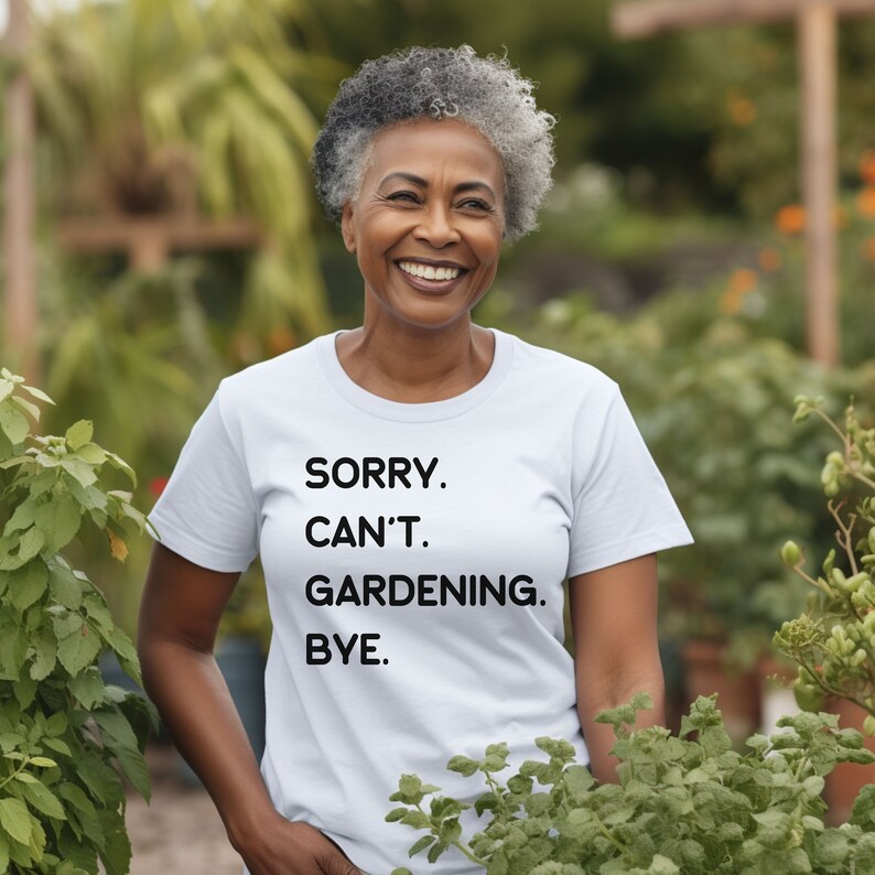 K&ouml;nnte beinhalten: Eine Frau tr&auml;gt ein wei&szlig;es T-Shirt mit dem Text "SORRY. CAN'T. GARDENING. BYE." in Schwarz gedruckt. Sie steht in einem Garten mit gr&uuml;nen Pflanzen.