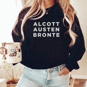 May include: Black sweatshirt with white text that reads "ALCOTT AUSTEN BRONTE". A person is wearing the sweatshirt and holding a floral mug.