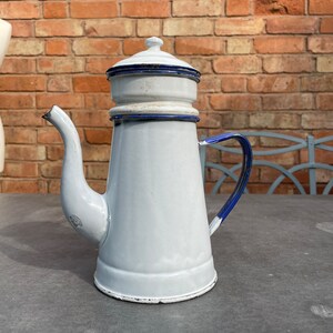 May include: White enamel coffee pot with a blue trim and a lid. The pot has a spout and a handle.