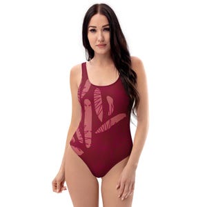 Pode incluir: Um fato de banho de uma peça em bordô escuro, com um design abstrato repetido em um tom mais claro de bordô. O fato de banho tem um decote redondo e uma silhueta clássica, adequado para nadar ou tomar sol.