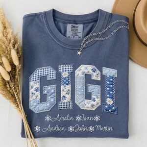 Puede incluir: Una camiseta azul Comfort Colors con el nombre "GIGI" en letras de patchwork. Debajo del nombre hay nombres adicionales. Un collar plateado con un colgante de estrella descansa sobre la camisa. Un sombrero beige y flores secas están en el fondo.