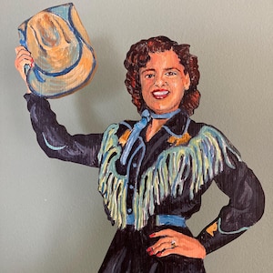 May include: A hand-painted wooden cutout of a person with dark wavy hair, wearing a black long-sleeved shirt with light blue fringe and a matching blue neck scarf. The figure holds a tan cowboy hat aloft in one hand and wears a ring on the other.