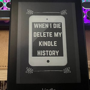 May include: A black Kindle e-reader displaying the text "WHEN I DIE DELETE MY KINDLE HISTORY" in a white rectangle. The Kindle is on a desk with a keyboard in the background.