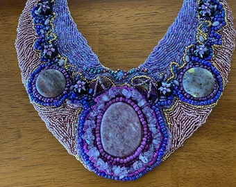May include: A purple and blue beaded necklace with a large oval-shaped stone in the centre. The necklace is decorated with smaller stones and beads in various shades of purple and blue.