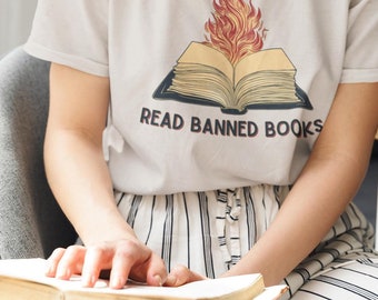 Puede incluir: Una persona con una camiseta blanca con el texto "READ BANNED BOOKS" y un gráfico de un libro abierto con llamas. La persona lleva gafas y sostiene un libro. La persona está sentada en una silla gris.