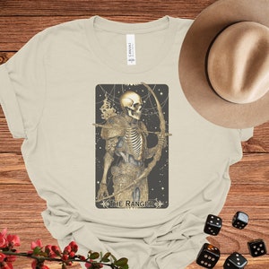 May include: A beige t-shirt with a graphic of a skeletal figure holding a bow and arrow. The graphic is surrounded by a black frame and the text "The Ranger" is printed below the image.