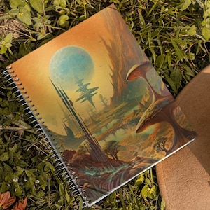 May include: A brown leather cowboy hat and a spiral bound notebook with a colorful illustration of a fantasy landscape on the cover are lying on a bed of green grass.