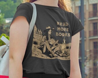 May include: A woman wearing a black t-shirt with a graphic of a woman reading a book by a campfire. The text on the shirt reads "Read More Books".