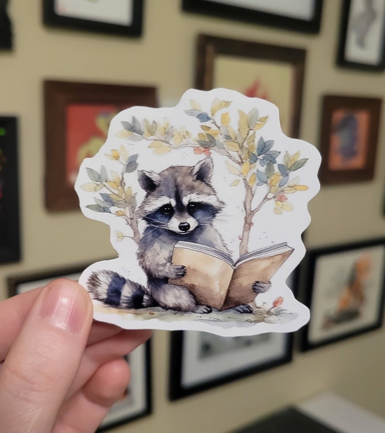 May include: A watercolor sticker of a raccoon reading a book. The raccoon is sitting on a branch with leaves and berries. The book is open and the raccoon is holding it with its paws.