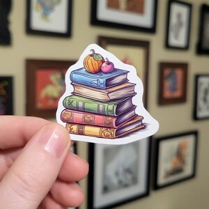 May include: A stack of five colourful books with a small orange pumpkin and a red apple on top. The books have different designs and are stacked on top of each other.