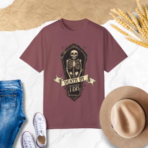 May include: A maroon t-shirt with a graphic of a skeleton in a coffin. The text "Death by TBR" is on a banner above the coffin.