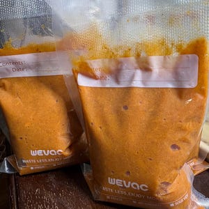 Può includere: Due sacchetti trasparenti per congelatore pieni di un prodotto alimentare arancione. I sacchetti hanno etichette bianche per il contenuto e la data. I sacchetti sono etichettati "WEVAC" e "WASTE LESS, ENJOY MORE".