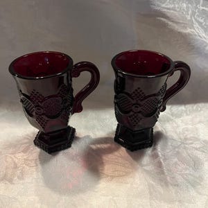 May include: Two ruby red glass mugs with ornate embossed patterns. Each mug has a handle and a hexagonal base. The mugs are displayed on a white patterned surface.
