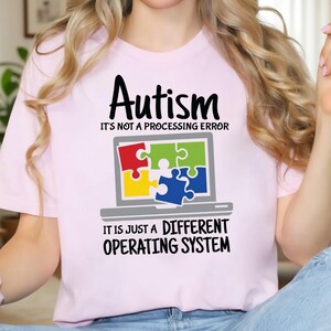 May include: A light pink t-shirt with the word "Autism" in black script above a laptop graphic. The laptop screen displays puzzle pieces. The text below the graphic reads "It's not a processing error. It is just a different operating system."