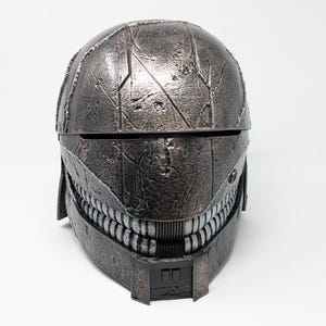 Op de afbeelding: Een donkergrijze helm met een getextureerd oppervlak en een gedetailleerd ontwerp. De helm heeft een vizier en een onderkaakstuk met een rij tanden.