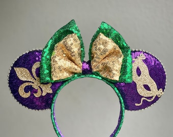 Puede incluir: Orejas de Minnie Mouse de lentejuelas moradas y verdes con un lazo dorado y un diseño de flor de lis dorada en una oreja y un diseño de máscara dorada en la otra oreja.