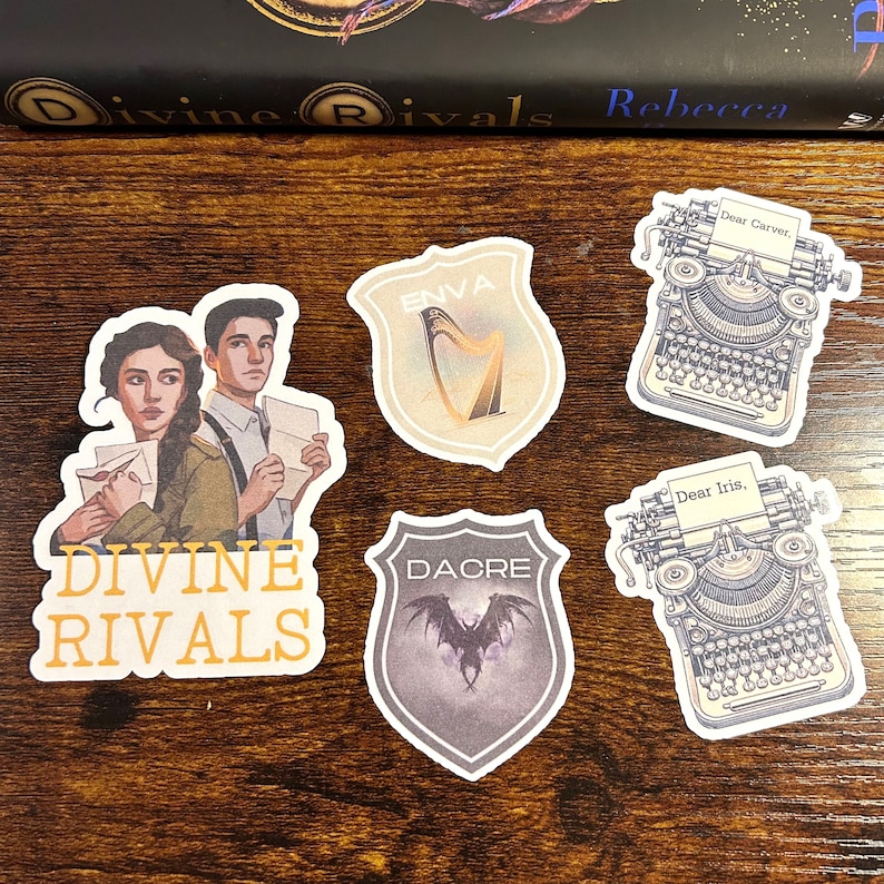 May include: Four stickers featuring characters and symbols from the book "Divine Rivals" by Rebecca. The stickers feature a harp, a bat, a typewriter with the text "Dear Iris" and a typewriter with the text "Dear Carver".