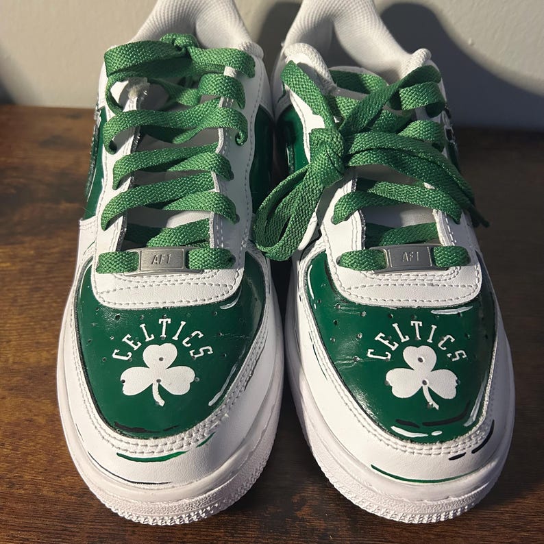 May include: Pair of white and green sneakers with green laces. The sneakers feature the word "CELTICS" and a shamrock logo on the toe. The shoes have a white sole and green accents.