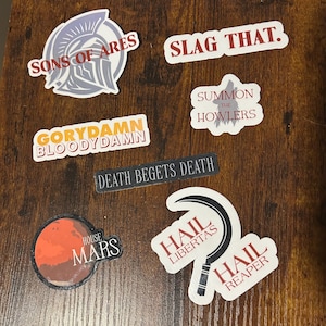 May include: A collection of stickers with various phrases and graphics. The stickers include text such as "SONS OF ARES," "SLAG THAT," "SUMMON THE HOWLERS," "GORYDAMN BLOODYDAMN," "DEATH BEGETS DEATH," "HOUSE MARS," "HAIL LIBERTAS," and "HAIL REAPER."