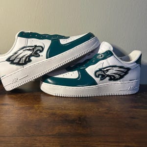 May include: White and teal sneakers with a Philadelphia Eagles logo on the side. The shoes have a white sole and teal accents. The logo is a detailed illustration of an eagle's head.