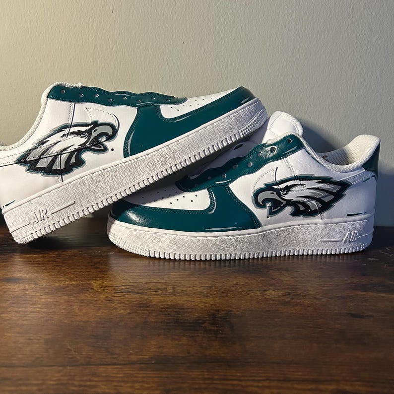 May include: White and teal sneakers with a Philadelphia Eagles logo on the side. The shoes have a white sole and teal accents. The logo is a detailed illustration of an eagle's head.