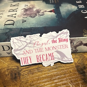 May include: A white sticker with a black and white illustration of a tree branch with a crow perched on it. The text "The girl, the King... And the Monster They Became" is written in pink on the sticker.