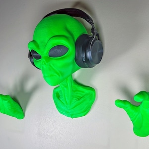 Puede incluir: Un busto de extraterrestre verde con auriculares puestos, con una expresión seria. El extraterrestre tiene dos manos extendidas, una a cada lado del busto.
