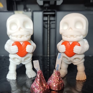 Puede incluir: Dos figuritas de esqueleto blancas sosteniendo objetos rojos en forma de corazón. Las figuritas están de pie sobre una superficie negra con dos besos de chocolate envueltos en papel de aluminio rosa delante de ellas.