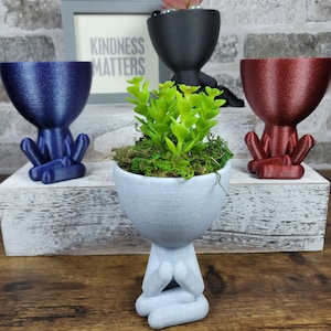 Puede incluir: Tres macetas de cerámica con forma de persona sentada en posición de meditación. Las macetas son azules, rojas y grises. La maceta gris tiene una pequeña planta verde en su interior. El letrero detrás de las macetas dice "La amabilidad importa".